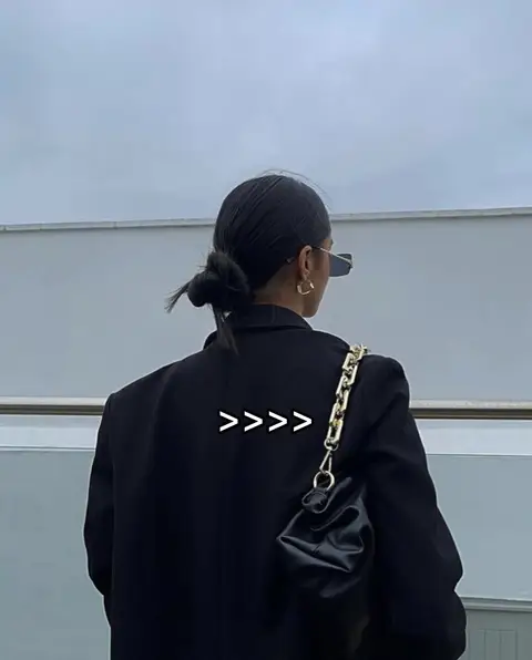 A woman with dark hair styled in a bun is seen from the back, wearing sunglasses and a black blazer. She is holding a black bag with a metallic chain strap on her right shoulder against an overcast sky and a white wall. The text overlay on the jacket back reads as several right-pointing arrows.