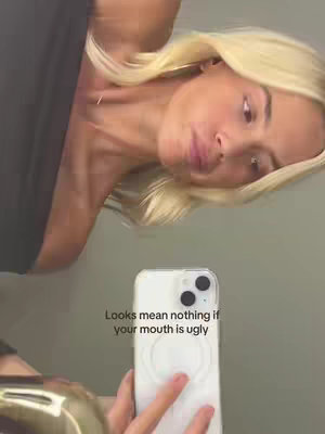 A selfie video shows a woman holding her phone in front of a mirror. The woman has long blonde hair, is wearing a dark top, and appears to be looking down at her phone as she records. Text is overlaid on the image.
