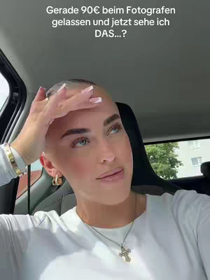 A woman with a shaved head is inside a car, looking up and to the right with a thoughtful expression. Text in German is overlaid at the top. She wears a white shirt, a necklace with a cross pendant, and gold hoop earrings. Her hand is raised to her head.