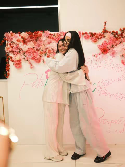 The cover image shows two young women embracing each other in front of a wall. The background is decorated with floral arrangements in shades of pink and red. The women are both smiling, and their arms are wrapped around each other. One woman is wearing glasses and a head covering, while the other has dark hair. The scene is warmly lit, creating a sense of intimacy.