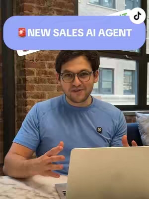 A man with glasses sits at a table with a laptop, facing the camera. He has short dark hair and is wearing a blue t-shirt. Above him, a blue speech bubble reads 'NEW SALES AI AGENT'. The background includes a brick wall and a window.