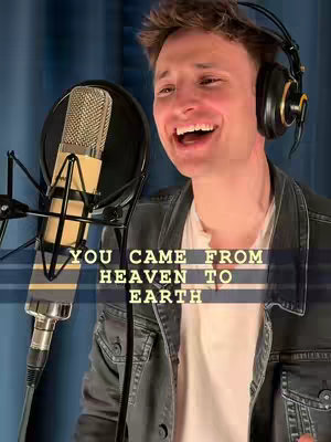 The image is a medium shot of a smiling man singing into a microphone. He wears a black jacket over a white shirt, and headphones are on his head. A microphone and pop filter are in front of him. Text overlays the image.