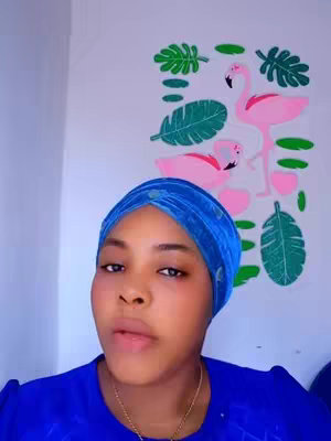 A woman is centered in the frame, looking towards the viewer. She wears a vibrant blue headscarf and a matching blue top, complemented by a gold necklace. Behind her, a wall is adorned with a painted mural featuring pink flamingos and green tropical leaves. The composition is simple, with the woman as the main subject, framed against the colorful background.
