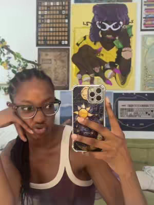 A young Black woman with glasses takes a selfie in a room. Behind her are colorful posters on the wall. She is holding her phone up in front of her face. Her expression is a slight frown with her lips forming a pout.