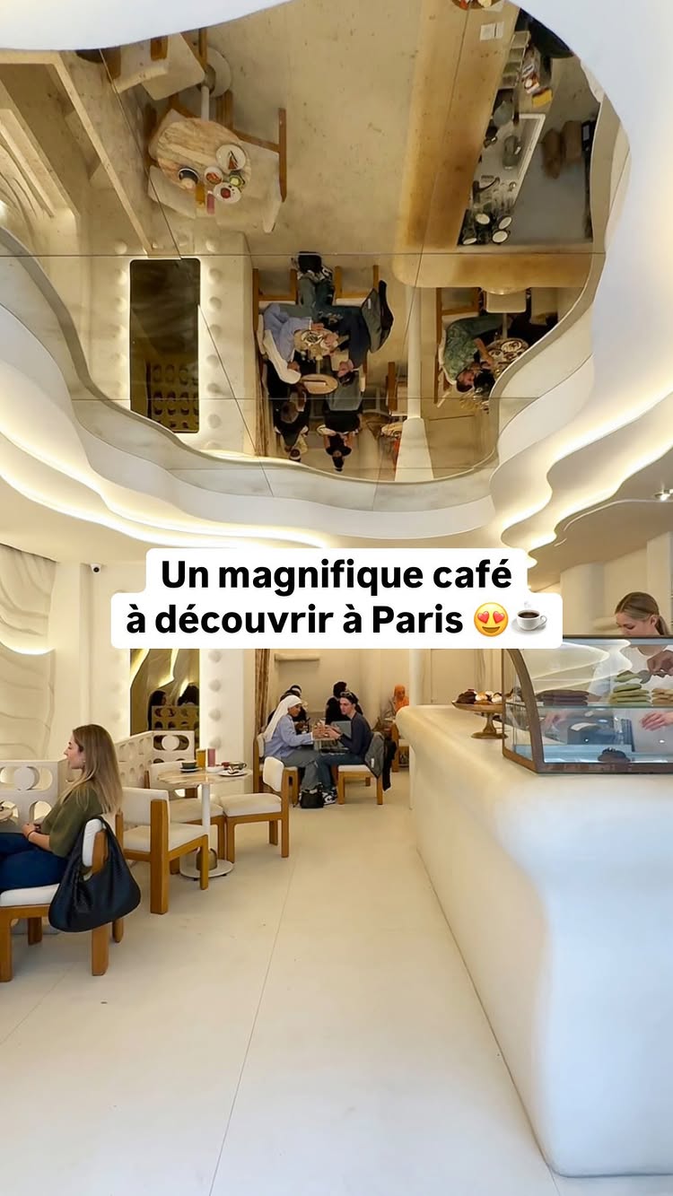 The cover image shows the interior of a modern coffee shop. The space is brightly lit and primarily white, with wavy, architectural elements defining the ceiling and walls. Tables and chairs are arranged for customers. In the foreground, a woman is seated. The image includes a mirrored ceiling, reflecting the shop's interior and enhancing the sense of space. A text overlay states, "Un magnifique café à découvrir à Paris" (A magnificent café to discover in Paris).