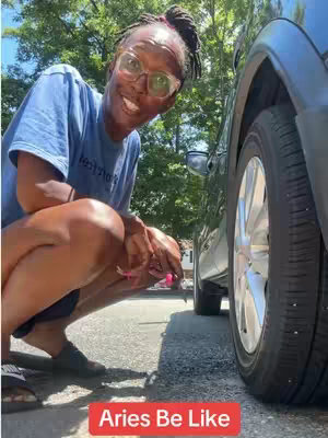 A woman is squatting on the pavement, next to a car tire, looking at the camera and smiling. The background has trees visible.