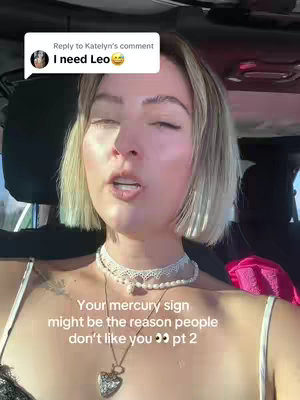 A woman with short blonde hair is positioned inside what appears to be a vehicle. The background is slightly blurred, suggesting movement or a shallow depth of field. She is wearing a white top and two necklaces. The image has a natural lighting, likely from daylight.