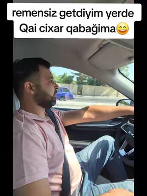A man driving a car, viewed from inside the vehicle. The man is centered and slightly to the left, steering the car and looking forward. The background shows a typical road scene with other cars and buildings visible through the windows. He wears a pink shirt and jeans. The image is taken during daylight hours.
