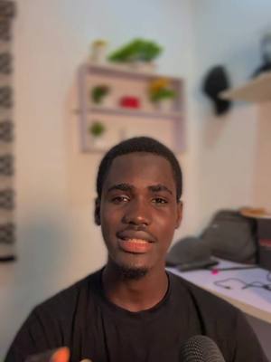 A medium shot shows a young Black man looking directly at the camera with a neutral expression. He has short, dark hair and a slight smile. His skin tone is warm. He wears a black shirt, and a microphone is visible, suggesting he might be recording. The background is softly blurred, showing a light-colored wall and a shelf with decorative items. The composition is well-lit and focused on the man.