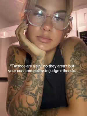A woman with blonde hair and glasses looks towards the camera pensively, resting her chin on her hand. She has numerous tattoos visible on her arms. Overlayed text provides a statement against judging others.