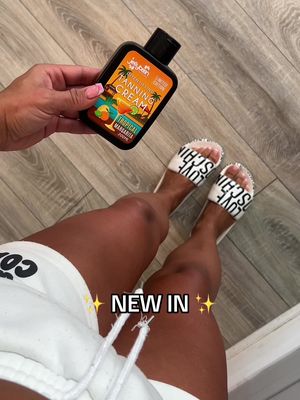 The cover image is a photograph featuring a person's legs and arms, highlighting a beauty or skincare product. The person is holding a black bottle of tanning cream with a colorful label that reads 'Tanning Cream Tropical Margarita' and shows an illustration of a tropical scene. The person is wearing white sandals with the word 'LOVE' repeated and white shorts. The legs are tanned with a wooden floor in the background. The overlay text reads 'NEW IN' with star emojis on both sides.