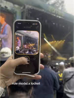 A vertical image shows a person holding a phone, displaying a view of a concert stage. The phone is centered in the frame, with a portion of the person's hand and arm visible. The phone screen displays a photo or video, with an overlay of a stage and performance. In the background, out-of-focus figures and lighting suggest a large outdoor venue.