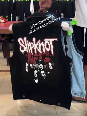 A person is holding a black vest with a Slipknot band logo on it. The vest is black with a graphic of the band members' faces in white and the band's name Slipknot in white and red letters. The background is blurry, but there are items and other vests visible.