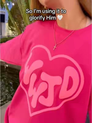 The image features a person wearing a bright pink sweatshirt with a large heart-shaped design. Inside the heart, the letters 'YTD' are visible. The person is also wearing a gold necklace with a cross pendant. The background is blurred, suggesting an outdoor setting with some greenery and possibly a fountain. The overall composition is well-lit and focuses on the upper body.