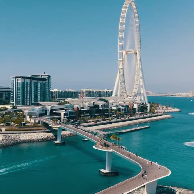 An aerial view shows a modern cityscape under a clear sky. A Ferris wheel stands prominently, with a curved bridge and water in the foreground. Buildings and coastal elements are visible.
