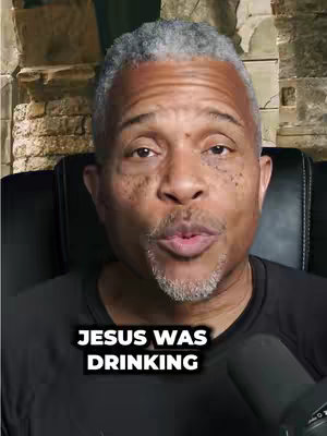 A medium shot of an older Black man with grey hair, wearing a black shirt, speaking directly into the assumed camera. Text overlay at the bottom reads, "JESUS WAS DRINKING", presented in a white sans-serif font with a black background. The background is a shallow depth of field showing a textural stone wall or fireplace.