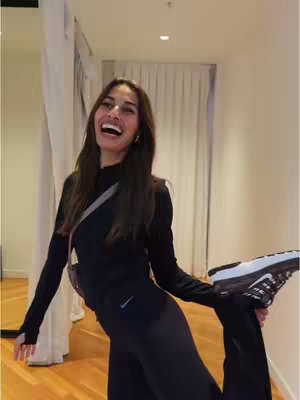 A woman in a black outfit is laughing and holding up a black and white Nike shoe in a hallway with cream-colored curtains and wooden floors. The lighting is soft and natural.