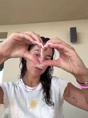 A young woman with wet, dark hair, is forming a heart shape with her hands in front of her face. She is wearing a white top with yellow trim and a small necklace. Behind her is a neutral-colored wall, with a small black rectangular object mounted on it.
