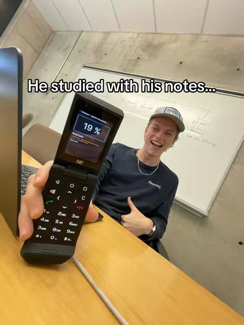 A young man smiling and giving a thumbs up while holding a flip phone with a screen displaying a percentage. The image is framed within an indoor setting, suggesting a casual or work-related context.