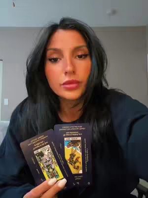 A woman with long, dark hair holds two tarot cards towards the camera. She is in a room with a light-colored wall and a glimpse of a couch. The image has a warm, natural lighting.
