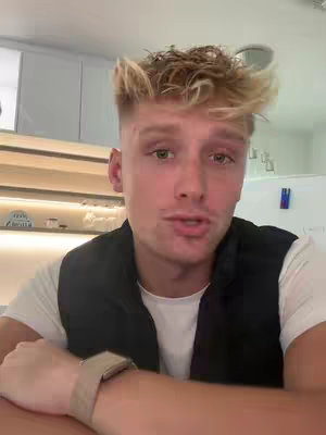A young man with short, styled blonde hair and a slight smile looks directly at the camera. He wears a white t-shirt under a black vest, and a watch is visible on his wrist. The background shows minimalist interior elements.