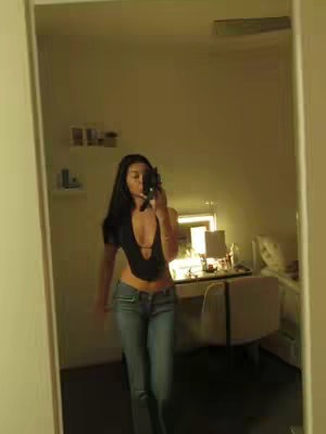 A full-length selfie in a mirror. The person is in the center wearing a black top with a low neckline, jeans, and a camera. The room is dimly lit background with a desk and a chair.