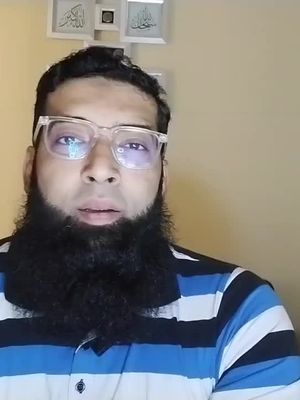 A man with glasses and a full, dark beard looks directly at the camera. He is wearing a striped shirt with vertical stripes of blue and white. The background is a plain, light beige wall with three framed Islamic calligraphy pieces.