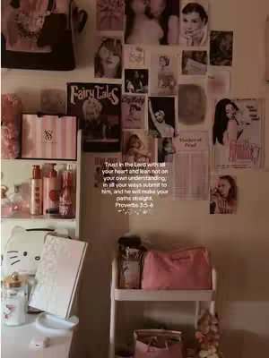 A room interior showcases a wall adorned with various printed images, including celebrity portraits, and magazine covers. Below the wall a section of shelves is visible with pink Victoria's Secret bags and bottles. The lighting is soft and warm.