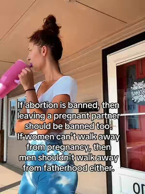 A woman stands outdoors holding a pink bottle. She is wearing a white top and blue and white patterned pants. Behind her is a building and a door sign
