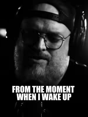 A grayscale close-up shot features a man with a beard, glasses, and a backwards baseball cap. He is wearing headphones and a microphone is positioned close to his mouth. Text in bold white font at the bottom reads 'FROM THE MOMENT WHEN I WAKE UP'.