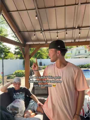 Two men are in an outdoor setting. One man is standing while pointing upwards. The other man is seated, with a clear bag on his lap. Text overlay reads, 'My bf said if this gets 50k likes he'll take me on vacation'.