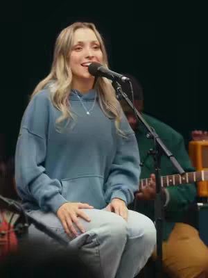 A woman with blonde hair is singing into a microphone, with a microphone stand in front of her. She is wearing a blue hoodie and light-colored jeans. A guitar and other bandmates are blurred in the background.
