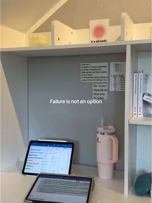 A well-lit desk setup with study materials. Two tablets with open documents sit on the desk. A pink water bottle and stationery are also visible. The background includes a white shelf with notebooks and a gray wall with sticky notes and motivational text: "Failure is not an option".