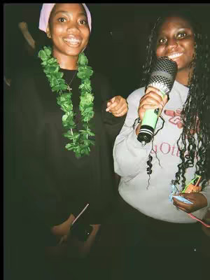 Two young women are posing, smiling, in a dimly lit setting that looks like a stage. The woman on the left is wearing a black sweater and a pink head covering, adorned with a green floral garland. The woman on the right is holding a microphone, and wearing a gray sweatshirt with a red, white and black logo.