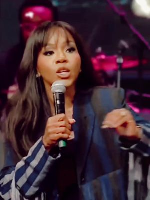A woman with dark hair and a microphone speaks passionately. She wears a blazer with blue and gray stripes, and the background is dimly lit with stage lighting.