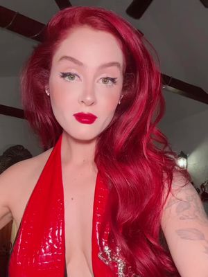 A woman with vibrant red hair and bold red lipstick gazes directly at the camera. She's wearing a red halter top that is glossy, and the background has ceiling fan blades. The lighting is even, highlighting her facial features.