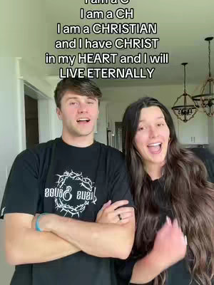 The image shows two people, a man and a woman, standing close together indoors. The man has short brown hair and is wearing a black t-shirt with a white design on it; he has his arms crossed. The woman has long dark hair and is wearing a black top, and she has her mouth open as if she is talking or singing. Text overlay in white letters at the top of the image reads, 'I am a C, I am a CH, I am a CHRISTIAN and I have CHRIST in my HEART and I will LIVE ETERNALLY'. The background includes a doorway and the corner of a room with a light above.