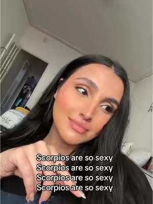 A woman is seen in a selfie-style video. The camera angle is from the upper body, showing her face and upper chest. She has long, dark hair. The background is a home environment, with glimpses of a door and other interior elements. The overlaid text reads: "Scorpios are so sexy" repeated multiple times.