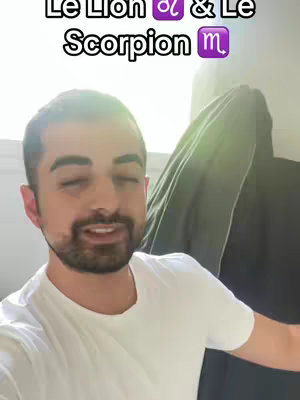 A man with short, dark hair and a beard is centered in the frame, looking towards the viewer and talking. Above him, text overlayed reads "Le Lion & Le Scorpion". The background appears to be a bright room with a dark, possibly gray, garment hanging from the top right of the frame.