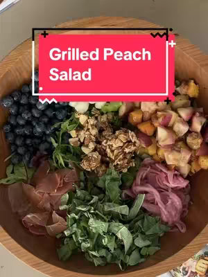 Overhead shot of a wooden bowl filled with a colorful salad. The salad is divided into sections or rows, including blueberries, a cluster of nuts, prosciutto slices, green leaves, pickled onions, and peach slices. A red rectangular text box with white text that reads 'Grilled Peach Salad' overlays the top of the image.