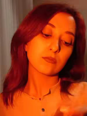 A portrait shot features a woman with reddish hair and fair skin, looking down with a slightly melancholic expression. The lighting is warm and orange, casting a gentle glow over her face and hair. The background is softly blurred, suggesting a close-up or intimate setting.