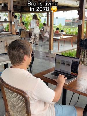 A man is seated at a wooden table in an outdoor setting, using a laptop. The scene appears to be a cafe or restaurant with other people in the background. Text overlay at the top left says 'Bro is living in 2078' followed by a sad emoji.