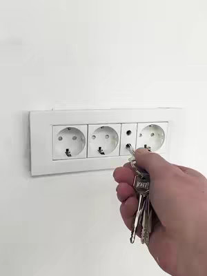 A hand holding a set of keys is inserting one into a Europlug socket on a white wall. There are three power outlets in a row, a bit dirty, with an extra hole in the middle for a pin. The white wall provides a minimalist background.