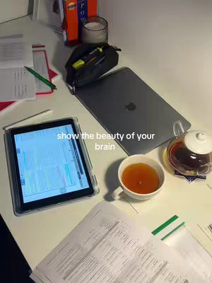 An overhead shot of a white desk with various items: a tablet displaying some coding on the screen, a laptop with a closed lid, a teacup, a tea pot, pens, and some books, all set on a white background. Text overlay reads 'show the beauty of your brain'.