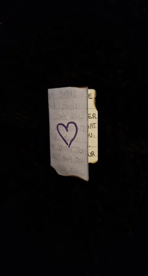 A close-up shot against a black background features a piece of paper with a handwritten note and a heart drawn on it. The paper is creased and has a burnt edge, suggesting it has been torn or damaged in some way. A dark, blurred background emphasizes the contrast.