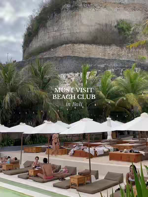 The image captures a scenic view of Palmilla Beach Club in Bali. The composition is focused on the beach club's amenities, including rows of sunbeds with umbrellas, set against a backdrop of a large, textured cliffside. The scene is bathed in natural light, creating a relaxing atmosphere. The color palette consists of neutral tones like beige, white, and brown, with accents of green from the surrounding palm trees and foliage. Several people are visible, enjoying the beach club setting.