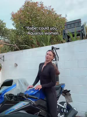 A woman in a black jumpsuit stands next to a blue and black motorcycle, with text overlayed. She is looking at the camera. A white wall and a tree form the background.