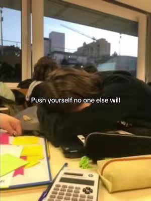 A person with their head down on a desk, possibly sleeping or resting, with a calculator, sticky notes, and a pencil case in the foreground. Outside a window, a cityscape and construction are visible.