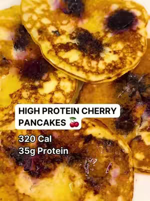 A close-up shot of several golden-brown pancakes topped with fresh cherries. The pancakes are stacked and glistening, with scattered pieces of cherries visible within. A white rectangular text box overlays the bottom of the image, displaying the text "320 Cal 35g Protein".