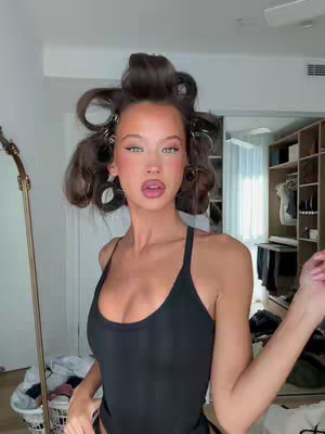 A woman with light skin and long brown hair is in a room with a neutral color scheme, wearing a black tank top. She has makeup, and hair curlers in her hair. The background suggests a bedroom, with a closet visible on the right and a mirror on the left. The woman is looking and slightly gesturing towards the camera.